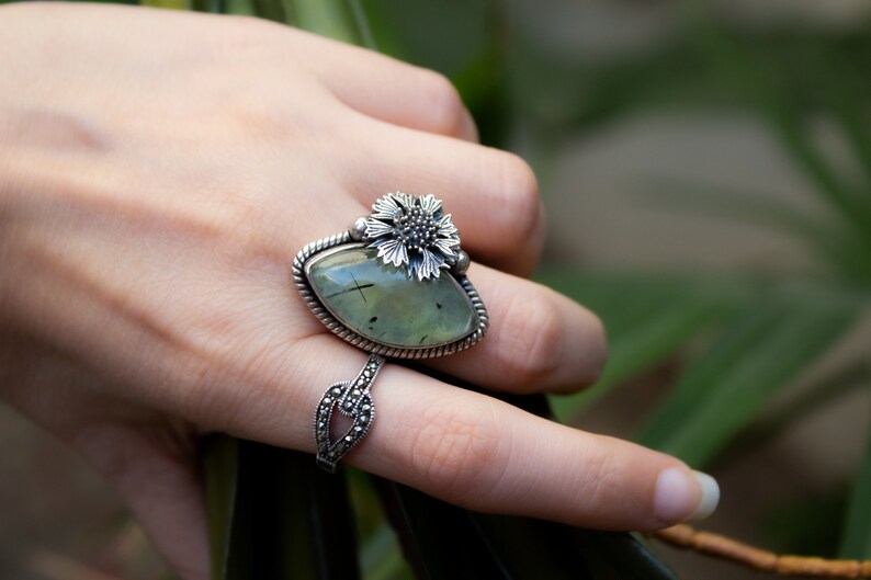 May include: A silver ring with a green gemstone and a flower design. The ring is on a finger.