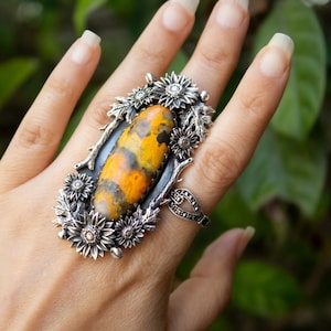 May include: A large statement ring featuring an elongated, oval-shaped bumblebee jasper stone with vibrant yellow, orange, and black patterns. The stone is set in a dark oxidised silver frame adorned with detailed metal sunflowers and leaves.