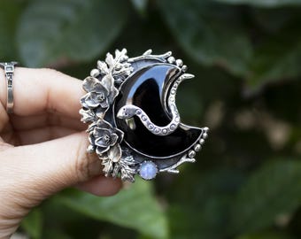 May include: A silver crescent moon pendant featuring a black onyx stone and a silver snake design. The pendant is surrounded by silver flowers and leaves.