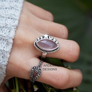 Op de afbeelding: Een zilveren ring met een roze rozenkwarts steen en een ster- en halvemaan ontwerp. De ring wordt gedragen op een vinger met een zilveren ring met een zwart stenen ontwerp. De tekst "ARDENT DESIGNS" is zichtbaar op de ring.