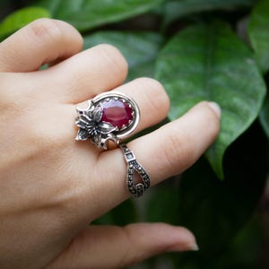 May include: Two silver rings are displayed on a finger. The first ring features a large, oval, deep red gemstone set in a decorative silver setting. The second ring is a smaller band with a repeating pattern of small silver details.