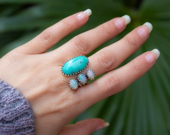 May include: A hand displays three rings with oval-shaped gemstones. The largest ring features a turquoise stone with a silver beaded border. Two smaller rings have opalescent stones. The rings are set against a blurred green background.