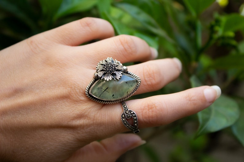 May include: A silver ring with a green gemstone and a flower design. The ring is on a person's finger.
