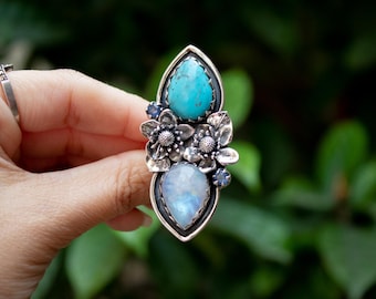 May include: A silver ring with two teardrop-shaped gemstones, one turquoise and one moonstone, surrounded by silver floral accents.