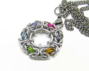 Puede incluir: Un collar con colgante de cota de malla plateada con un diseño circular. El colgante presenta un anillo de eslabones metálicos entrelazados, acentuado con eslabones coloridos en tonos rosa, morado, verde, azul y dorado. El collar tiene una cadena a juego.