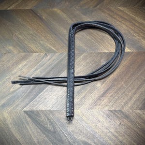 Studio-craft view of a Knotty Oso impact martinet handcrafted from black leather, featuring a hand-wrapped handle and multiple heavy straps.