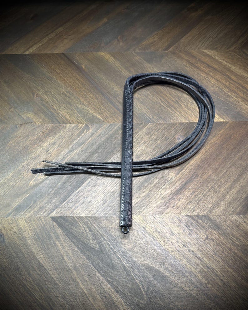 Studio-craft view of a Knotty Oso impact martinet handcrafted from black leather, featuring a hand-wrapped handle and multiple heavy straps.