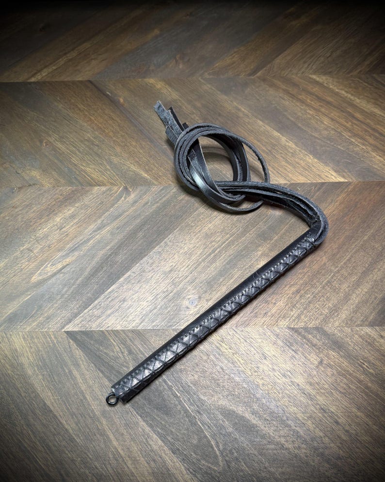 Hero-tilt view of a coiled black leather impact martinet with heavy-weight straps, showcasing structural material flexibility and construction density.
