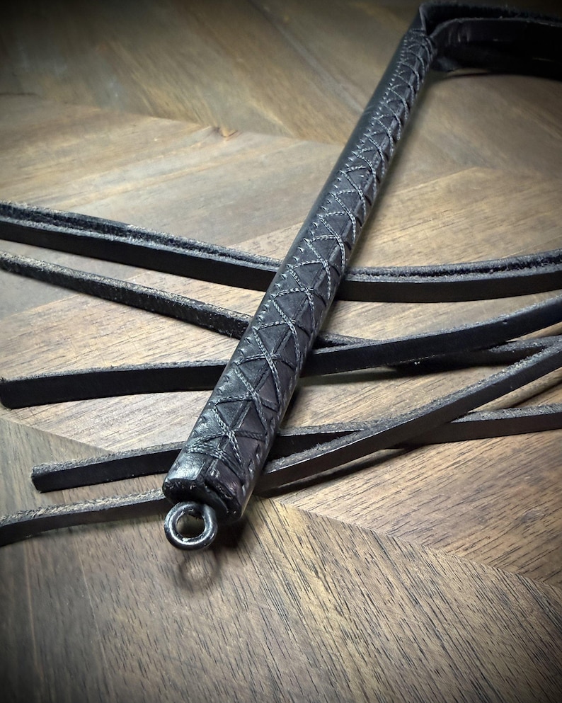 Detail-macro view of a black leather handle with a precision hand-sewn cross-stitch pattern, highlighting the integrated steel pommel ring.