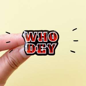 May include: A black and orange enamel pin with the words "WHO DEY" in a bold, white outline.