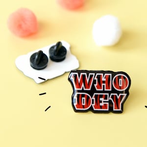 May include: A black and red enamel pin with the words "WHO DEY" in a tiger stripe pattern.