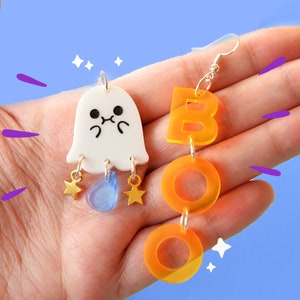 May include: A pair of Halloween-themed earrings. One earring features a white ghost with a cute face and blue and gold accents. The other earring features the word "BOO" in orange acrylic.