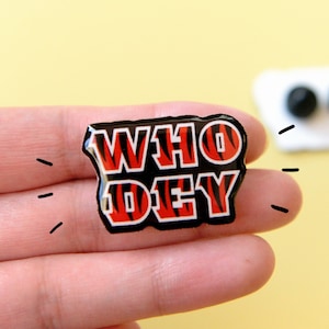 May include: A black and orange enamel pin with the words "WHO DEY" in white letters. The pin has a tiger stripe pattern.