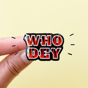 May include: A black and red enamel pin with a tiger stripe pattern. The pin says "WHO DEY" in white letters.
