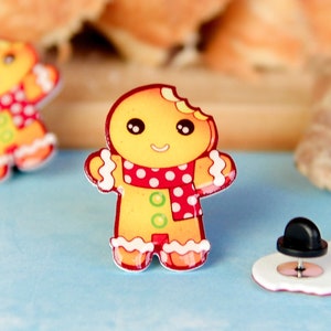 May include: Two gingerbread man pins with red and white accents. One gingerbread man is missing a bite. The pins are on a blue surface with a white gingerbread man pin in the background.