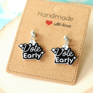 May include: A pair of silver stud earrings with black and white charms that say "Vote Early". The earrings are displayed on a wooden surface with a brown card that says "Handmade with love".