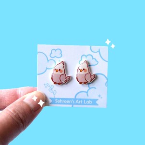 May include: A pair of pink bird-shaped earrings with orange beaks and white crests, displayed on a white card with cloud and leaf illustrations. The card has the text "Sehreen's Art Lab" on a light blue background.