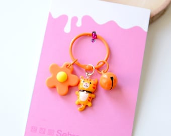 May include: Orange and yellow cat keychain with a flower and bell charm. The keychain is on a pink background with the text "SehrreensArtLab".