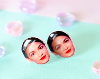 May include: A pair of stud earrings featuring a portrait of a woman with dark hair and red lipstick. The earrings are on a light blue surface with pink heart-shaped beads.