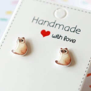 May include: A pair of silver stud earrings featuring a small, detailed illustration of a Siamese cat. The earrings are on a white card with the text "Handmade with love".