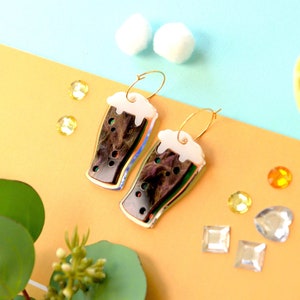 May include: Gold hoop earrings with a dark brown and iridescent glass charm shaped like a pint of beer with a white foam top.