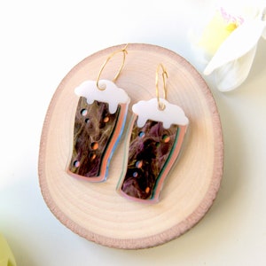May include: A pair of gold hoop earrings with a colorful, abstract design of a pint glass of beer. The glass is filled with a dark brown liquid and has a white foam head.