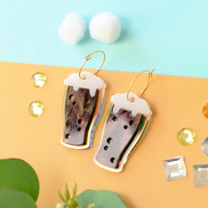 May include: A pair of gold hoop earrings with a brown and white glass beer mug charm. The mugs have a white foam top and a brown liquid inside.