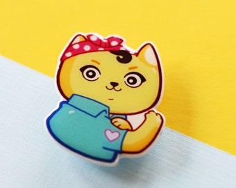 Puede incluir: Un pin de dibujos animados de gato amarillo con una diadema de lunares rojos y una camisa azul. El gato flexiona su brazo, con un corazón rosa en la camisa. El fondo es una división de amarillo y azul claro.