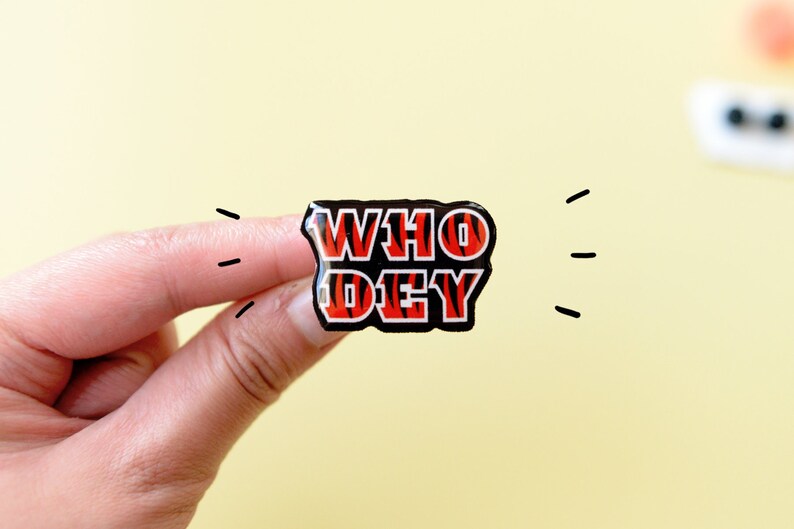 May include: A black and orange enamel pin with the words "WHO DEY" in a bold, white outline.