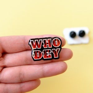 May include: A black and red enamel pin with the words "WHO DEY" in a tiger stripe pattern.