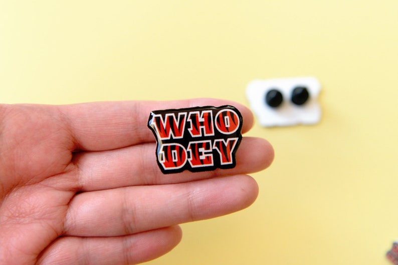 May include: A black and red enamel pin with the words "WHO DEY" in a tiger stripe pattern.