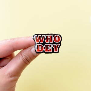 May include: A black and orange enamel pin with the words "WHO DEY" in white letters.