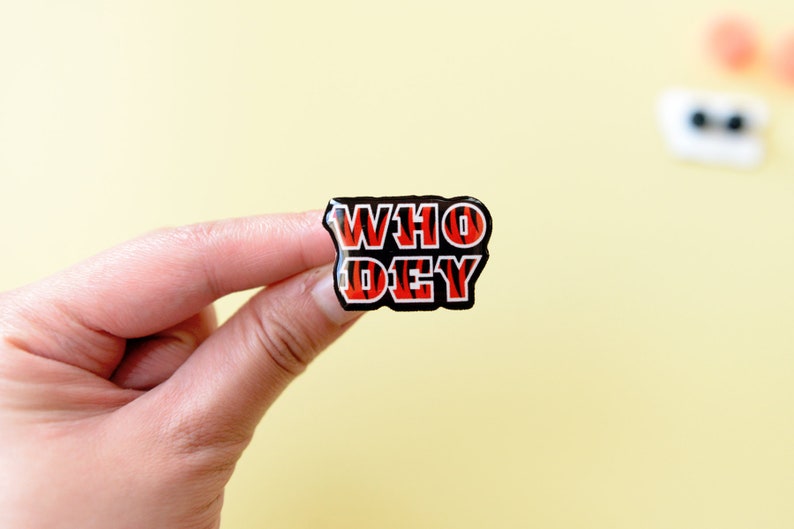 May include: A black and orange enamel pin with the words "WHO DEY" in white letters.