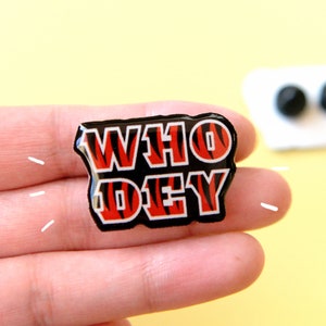 May include: A black and orange enamel pin with the words "WHO DEY" in white letters. The pin has a tiger stripe pattern.