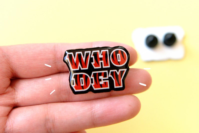 May include: A black and orange enamel pin with the words "WHO DEY" in white letters. The pin has a tiger stripe pattern.