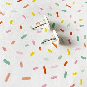 May include: A pair of white stud earrings with silver posts. The earrings are on a white background with a colorful confetti pattern.