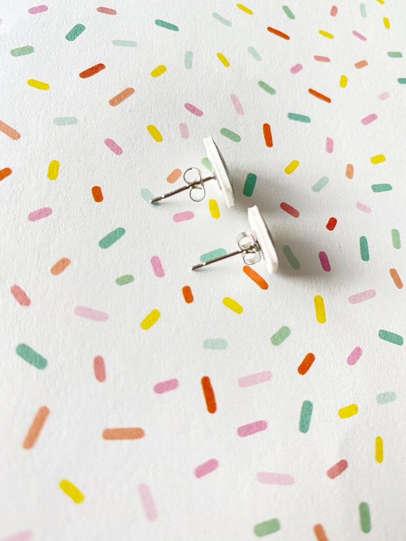 May include: A pair of white stud earrings with silver posts. The earrings are on a white background with a colorful confetti pattern.