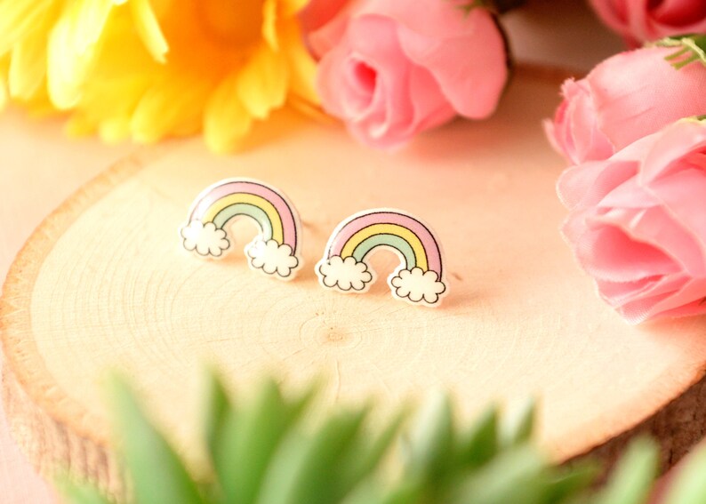 K&ouml;nnte beinhalten: Ein Paar Regenbogen-Ohrstecker. Jeder Ohrring hat ein farbenfrohes Regenbogen-Design mit pastellgelben, gr&uuml;nen und rosa B&ouml;gen und wei&szlig;en Wolkenakzenten. Die Ohrringe sind auf einer hellen Holzoberfl&auml;che mit Blumen im Hintergrund platziert.