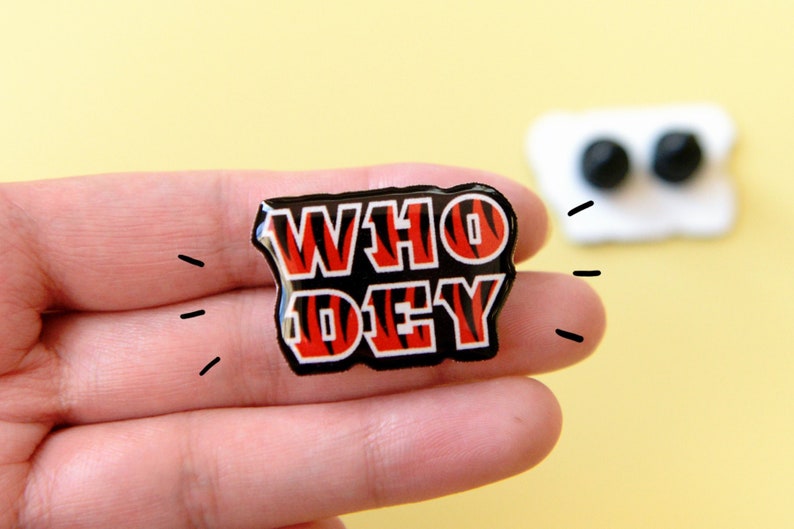 May include: A black and orange enamel pin with the words "WHO DEY" in white letters. The pin has a tiger stripe pattern.