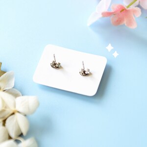 May include: A pair of silver-colored stud earrings with butterfly backs are displayed on a white card. The card is set against a light blue background, with decorative flowers in the corners.