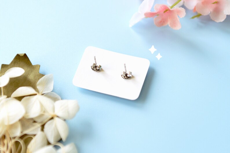 May include: A pair of silver-colored stud earrings with butterfly backs are displayed on a white card. The card is set against a light blue background, with decorative flowers in the corners.