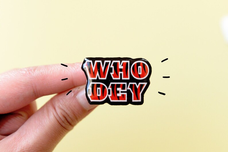 May include: A black and red enamel pin with a tiger stripe pattern. The pin says "WHO DEY" in white letters.