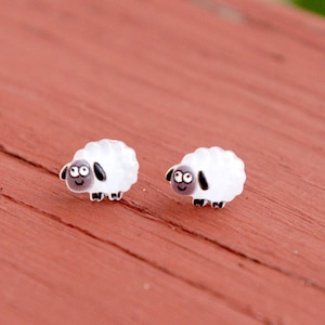 May include: A pair of whimsical sheep earrings. Each earring features a white, fluffy sheep with black accents on the ears, eyes, and hooves. The earrings are set against a reddish-brown wooden surface.