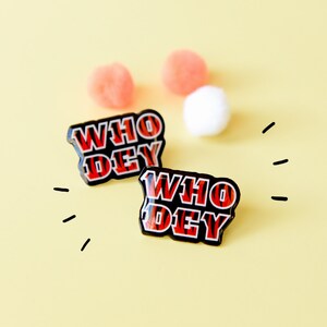May include: Two black enamel pins with the words "WHO DEY" in red and black striped lettering. The pins are on a yellow background with white and pink pom-poms.