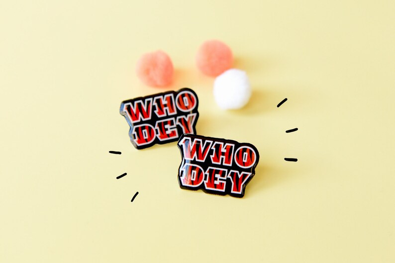 May include: Two black enamel pins with the words "WHO DEY" in red and black striped lettering. The pins are on a yellow background with white and pink pom-poms.