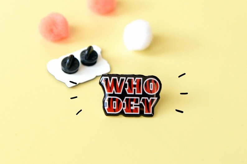 May include: A black and red enamel pin with the words "WHO DEY" in a tiger stripe pattern.