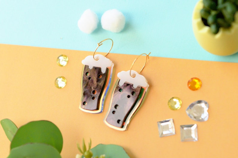 May include: A pair of gold hoop earrings with a brown and white glass beer mug charm. The mugs have a white foam top and a brown liquid inside.