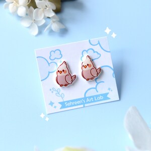 May include: A pair of bird-shaped stud earrings on a white card with blue cloud and star illustrations. The earrings are pink and white with a cartoon-like design. The card has the text "Sehreen's Art Lab".