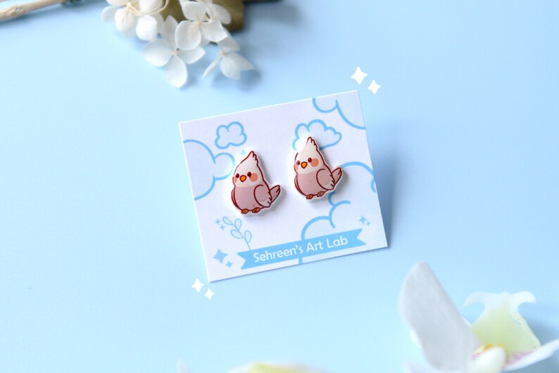 May include: A pair of bird-shaped stud earrings on a white card with blue cloud and star illustrations. The earrings are pink and white with a cartoon-like design. The card has the text "Sehreen's Art Lab".