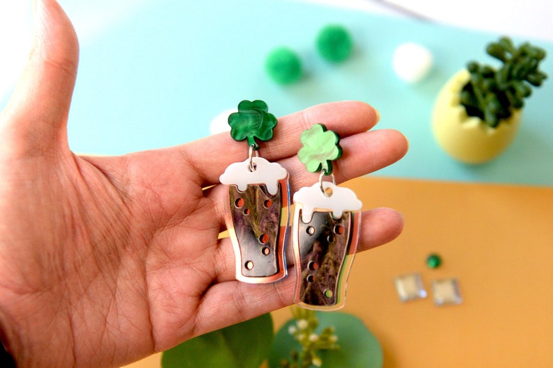 May include: A pair of dangle earrings featuring a clear glass beer mug with a white foam top and a green shamrock charm. The earrings are made of acrylic and have a silver finish.